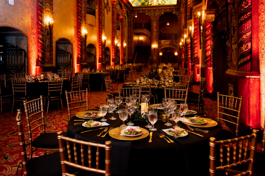A beautifully decorated ballroom at the Louisville Palace with plated salads, gold charger plates, gold chiavari chairs and black tablecloths in the foreground