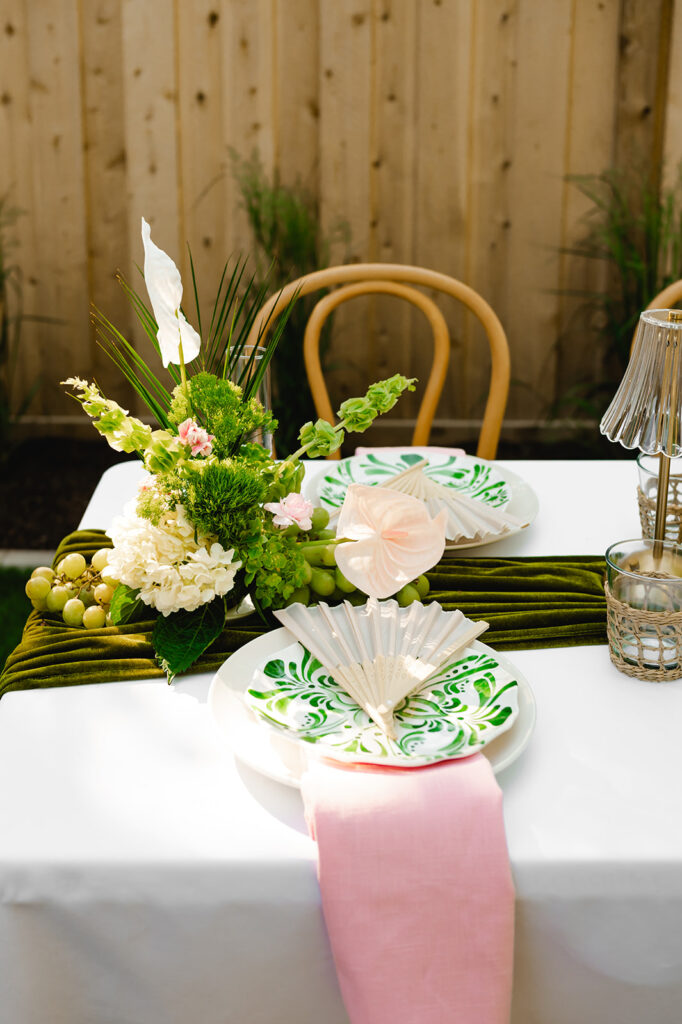 A tablescape for an event with the Palm Royale theme with a pink napkin, green patterned plate, and ivory fan at each place setting