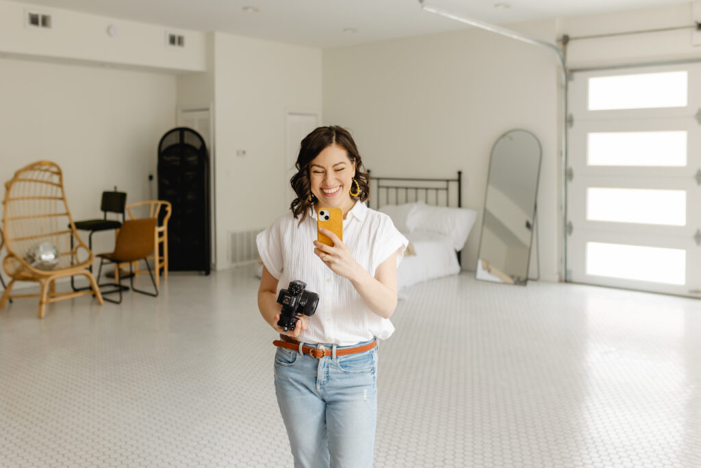 It's me, Johanna (your brand photographer) at Bright Studio Lou. I'm smiling and holding up my phone in one hand, my camera in the other. In the background, the wide open space of the studio is visible. On the left side, several chairs, a partition, and a disco ball are available for guests. Behind me is also a large mirror and a large garage style door with opaque panels that let in beautiful natural light.