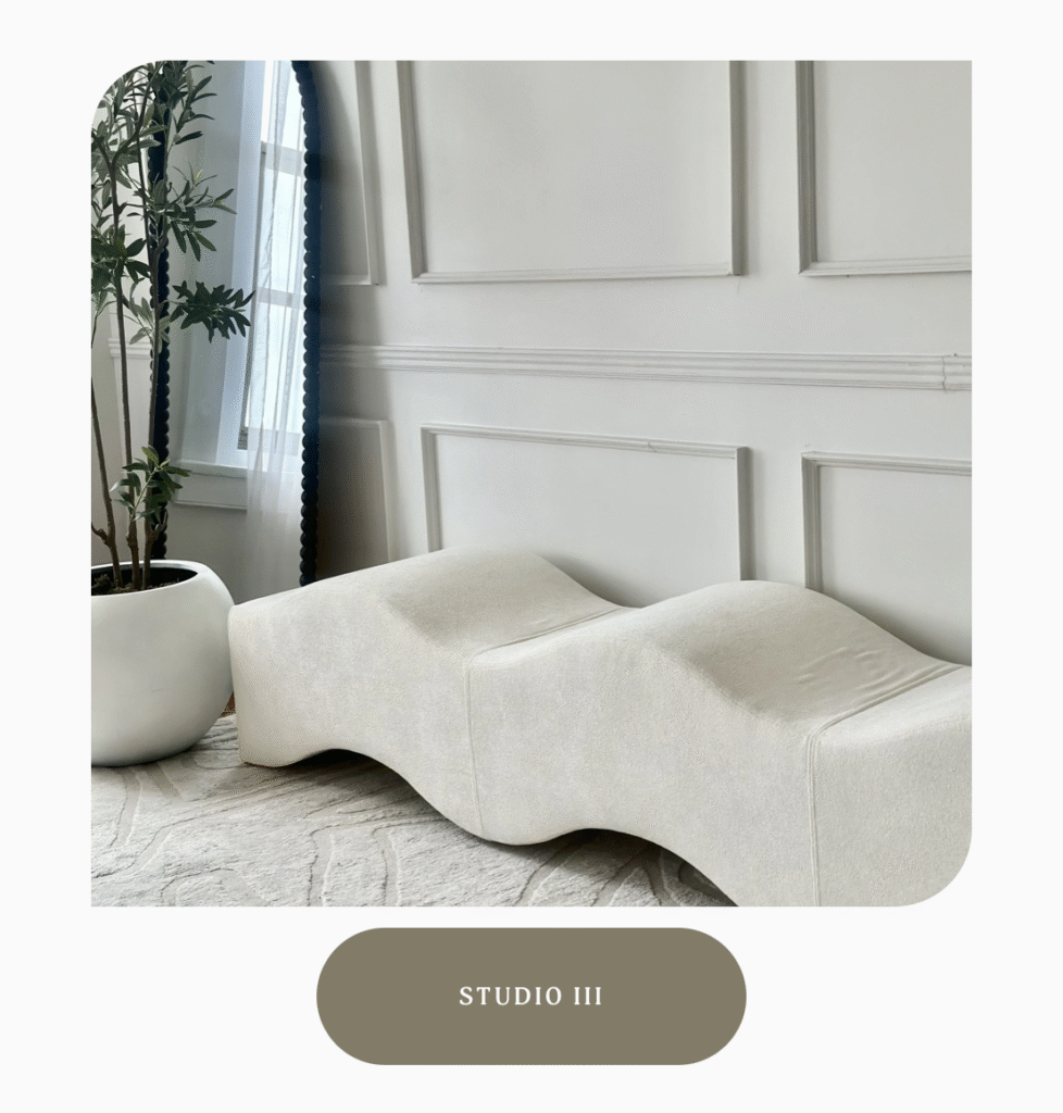 A neutral space with a white room and crown moulding, a wavy bench with white fabric, a faux tree in a white basin, and a large mirror.