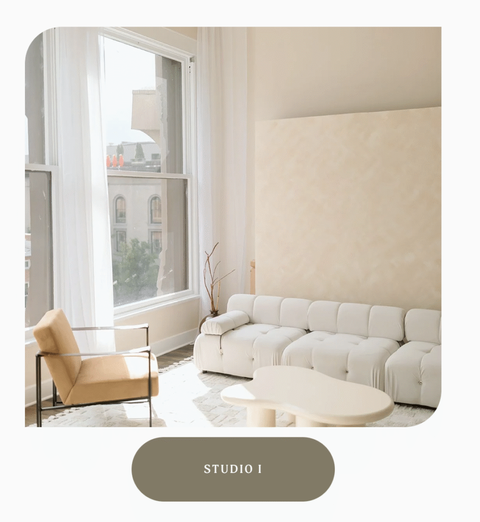 A natural light studio with large windows and sheer curtains overlooking Downtown Louisville. Inside the studio is a modern whtie couch, decorative wavy table, beige chair with metal armrests, and a textured, light beige rollable backdrop.