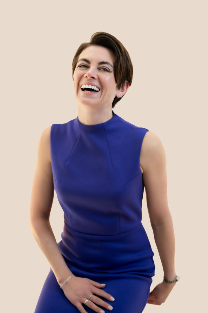 A professional woman with short hair, a radiant smile and wearing a  purple sleeveless dress laughs in this refreshingly candid headshot.