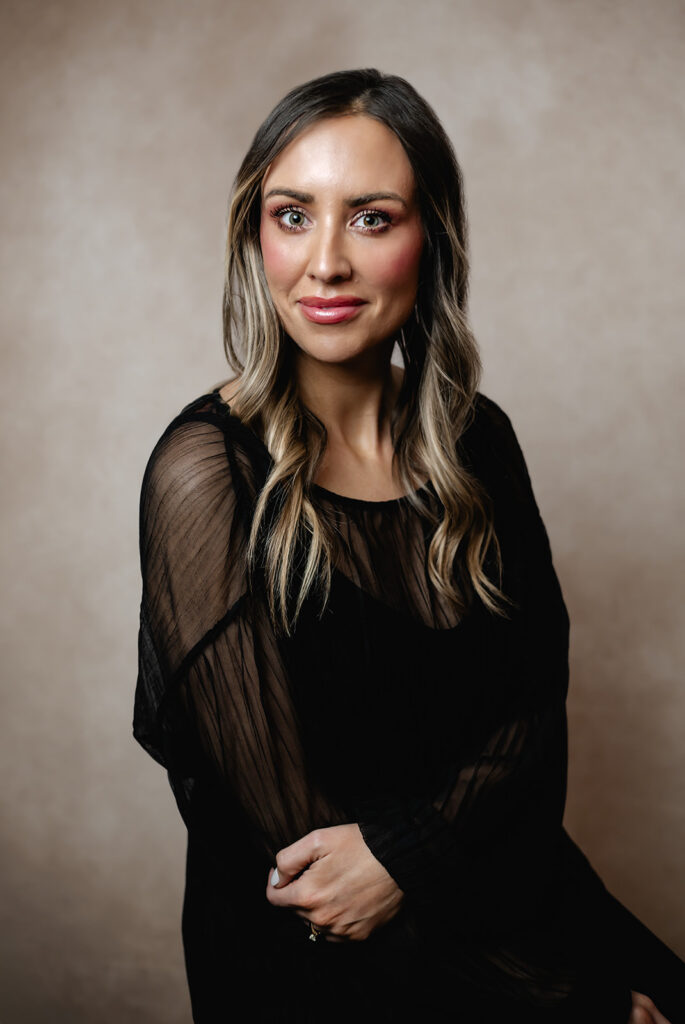 A beautiful makeup artist poses stoically. She wears a black blouse with sheer, textured sleeves, and her  romantic makeup look stands out against the textured beige backdrop.