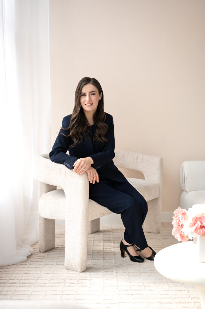 A wider cropped photo of a professional woman in a navy blue blazer and dress slacks sits with her hands resting on the white chair she is sitting in, and her ankles crossed in the Street View room of Loft Creative Studio.