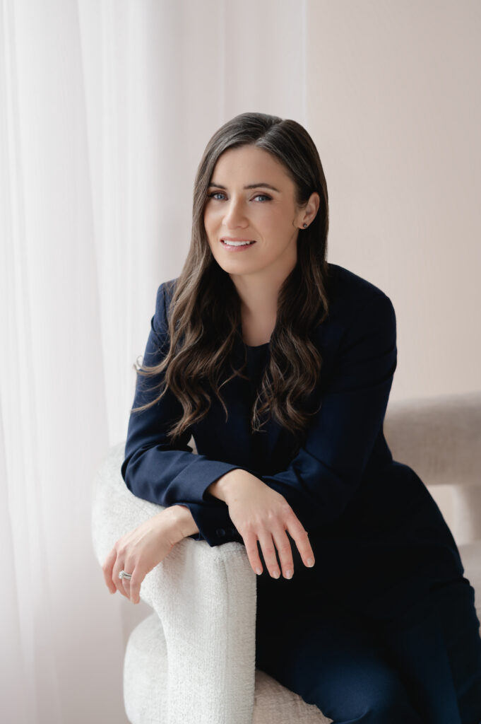 A professional woman in a navy blue blazer and dress slacks sits with her hands crossed in a relaxed pose in a bright, modern portrait studio.
