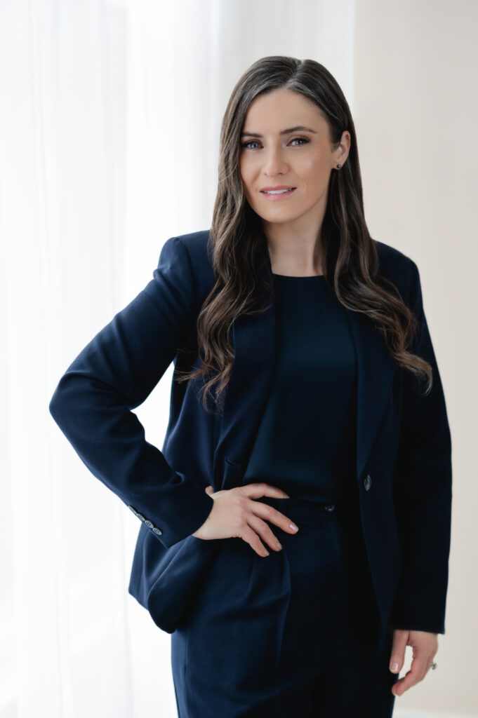 A standing portrait of a professional woman wearing a navy pantsuit. She smiles confidently with one arm on her hip, and the other relaxed. The natural light from the large window and sheer curtain behind her makes this portrait bright and welcoming.