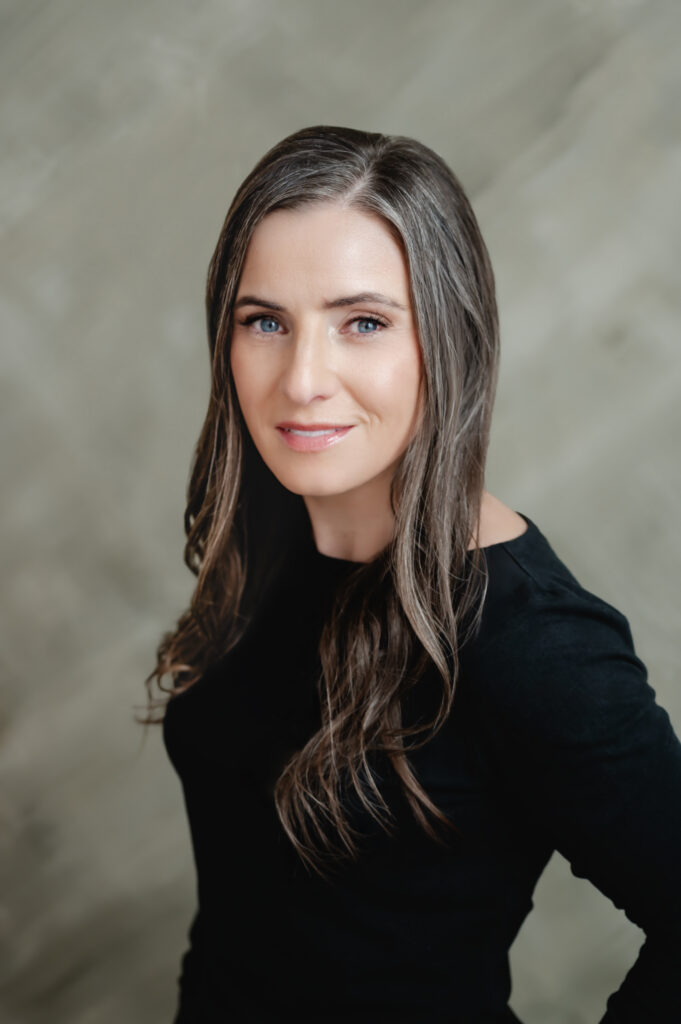 A closeup portrait of a woman in her 40s with long hair and a longsleeved black blouse against a textured green backdrop.