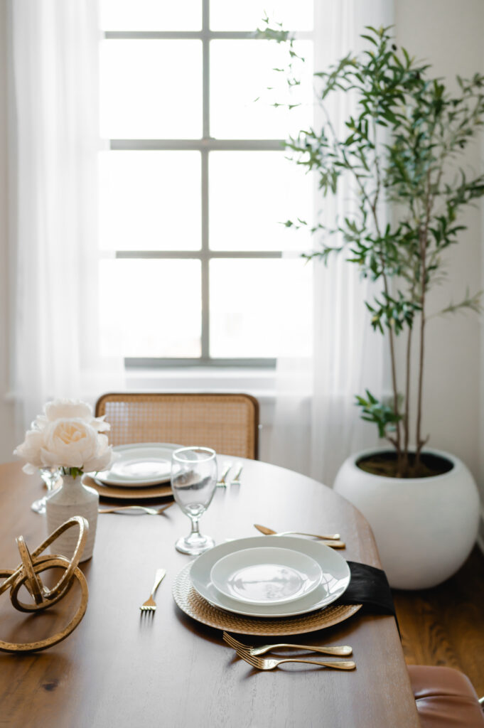 A styled scene in the River View Room of Loft Creative Studios. The neutral white walls with crown moulding are a blank canvas for your creative ideas. This scene features a long oval table, mid-century modern chairs with rattan backs and leather cushions, and a faux tree in a white basin. A large window with sheer curtains is in the background.