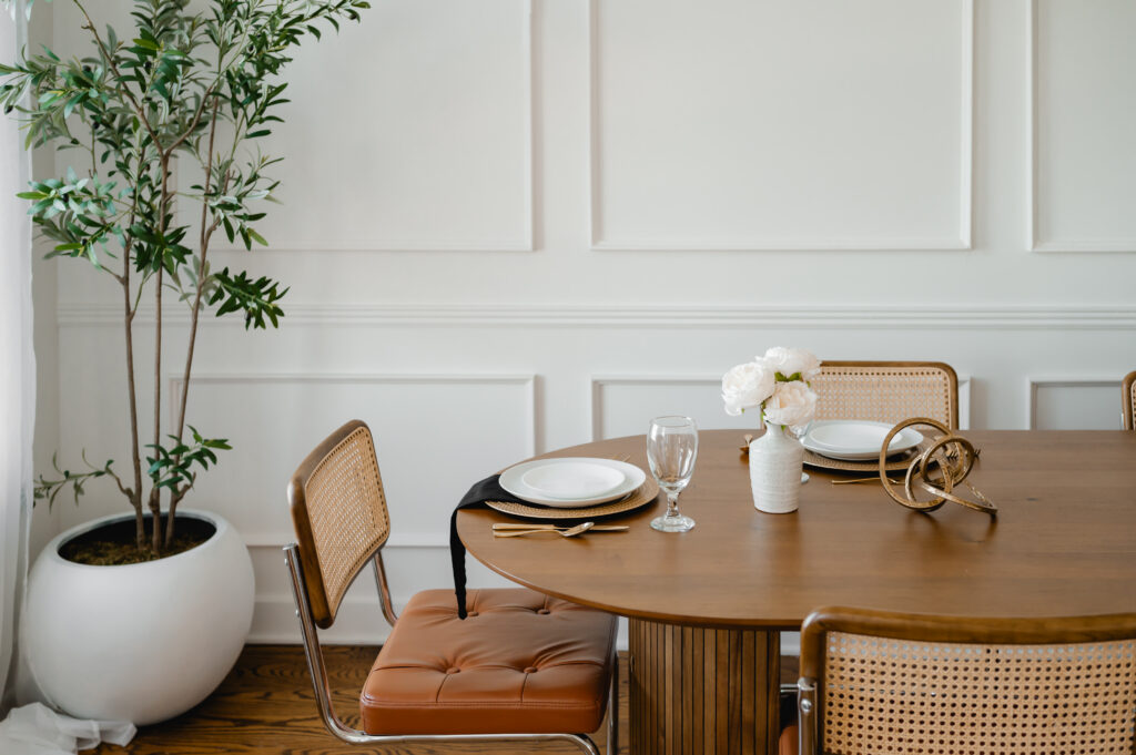 A styled scene in the River View Room of Loft Creative Studios. The neutral white walls with crown moulding are a blank canvas for your creative ideas. This scene features a long oval table, mid-century modern chairs with rattan backs and leather cushions, and a faux tree in a white basin.