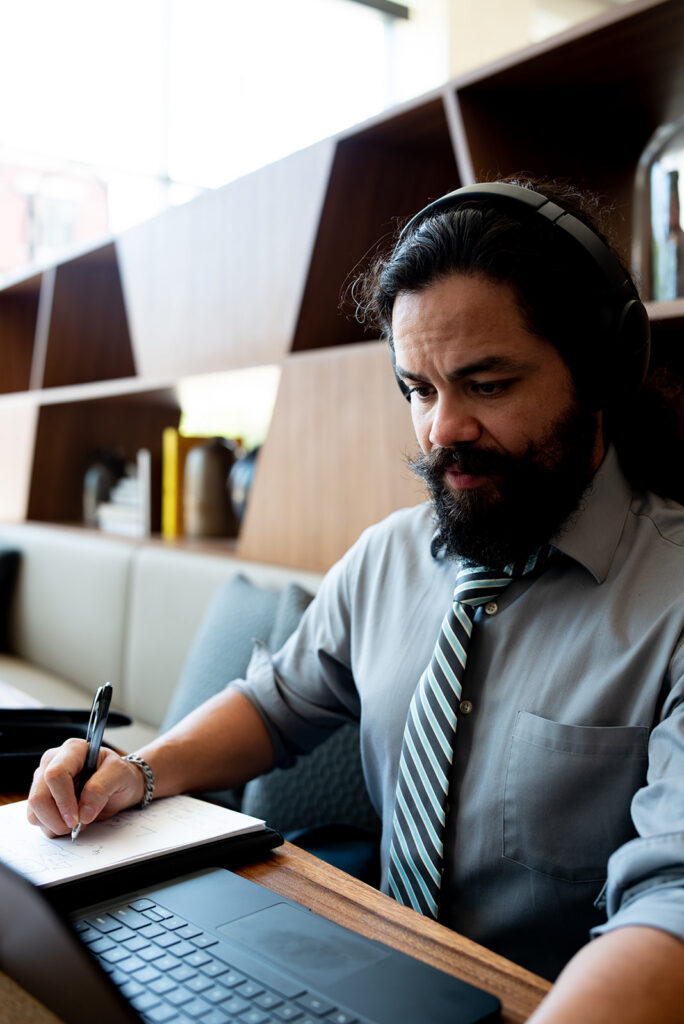 A handsome Hispanic interpreter with long curly hair pulled back and a thick beard wears a headset and looks at his computer as he takes notes on a notepad. He is in a booth in a mid-century inspired space.