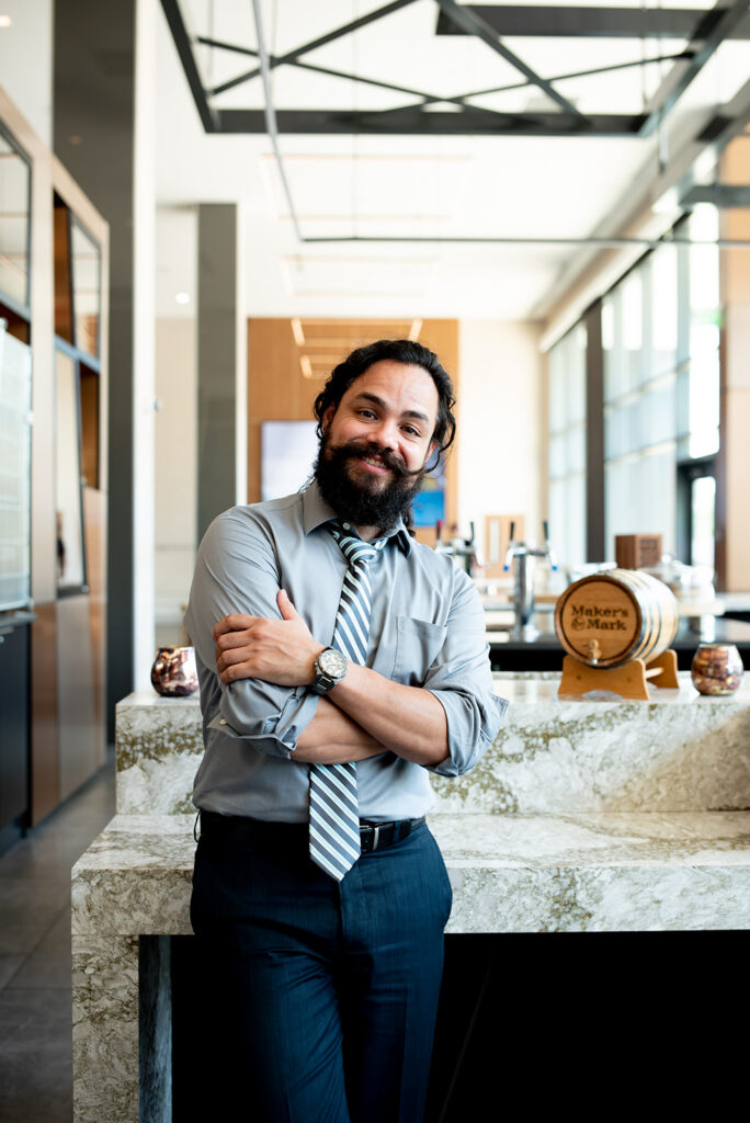 A handsome Hispanic professional with long curly hair pulled back and a thick beard leans against a marble counter in a naturally bright, open room. A small Maker's Mark barrel sits behind him, a nod to his time in Kentucky.