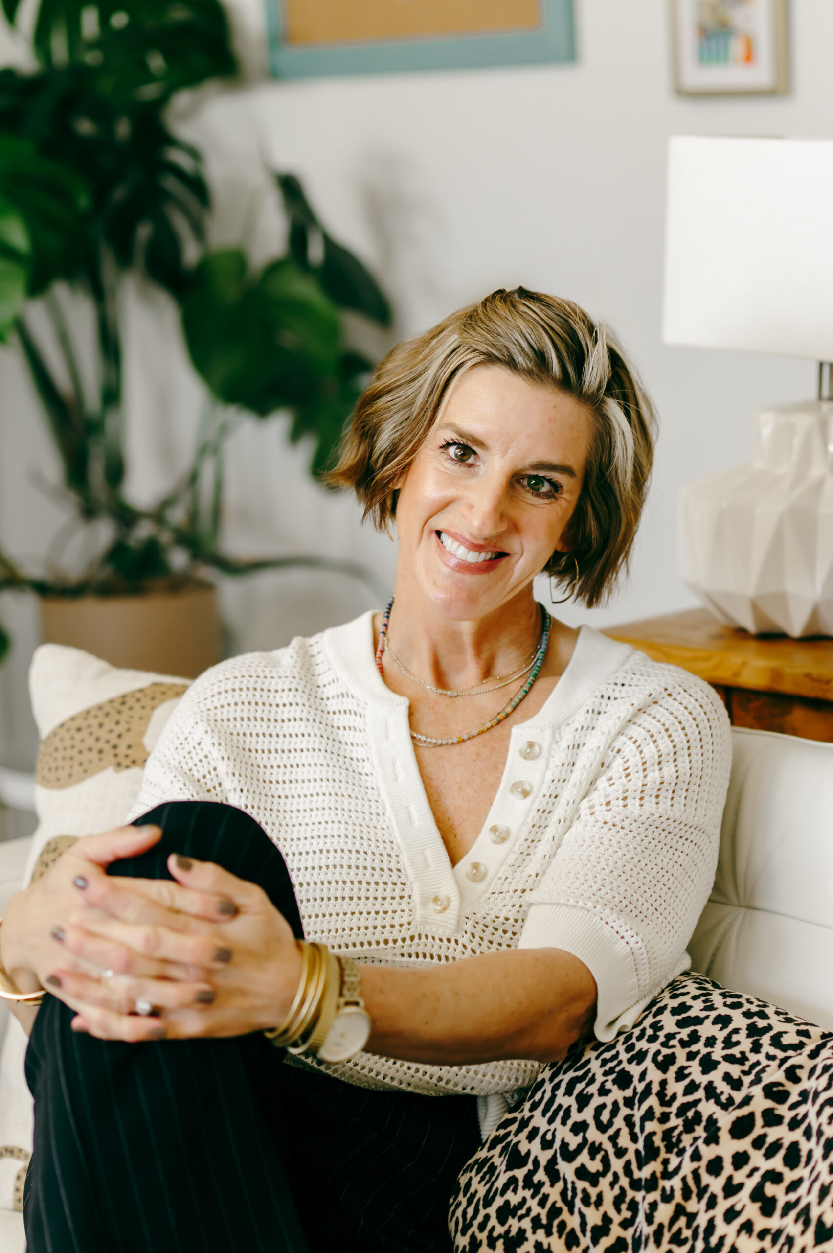 A Wardrobe Stylist Sits Relaxed on her office couch, wearing a comfortable woven ivory top and black linen pants.