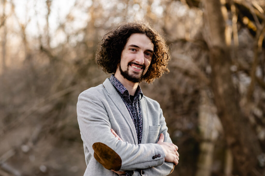The co-owner of a landscaping designer specializing in native pollinator gardens smiles for a natural light, environmental portrait outdoors. He wears a floral patterned dress shirt with a light grey blazer and stylish elbow patches.