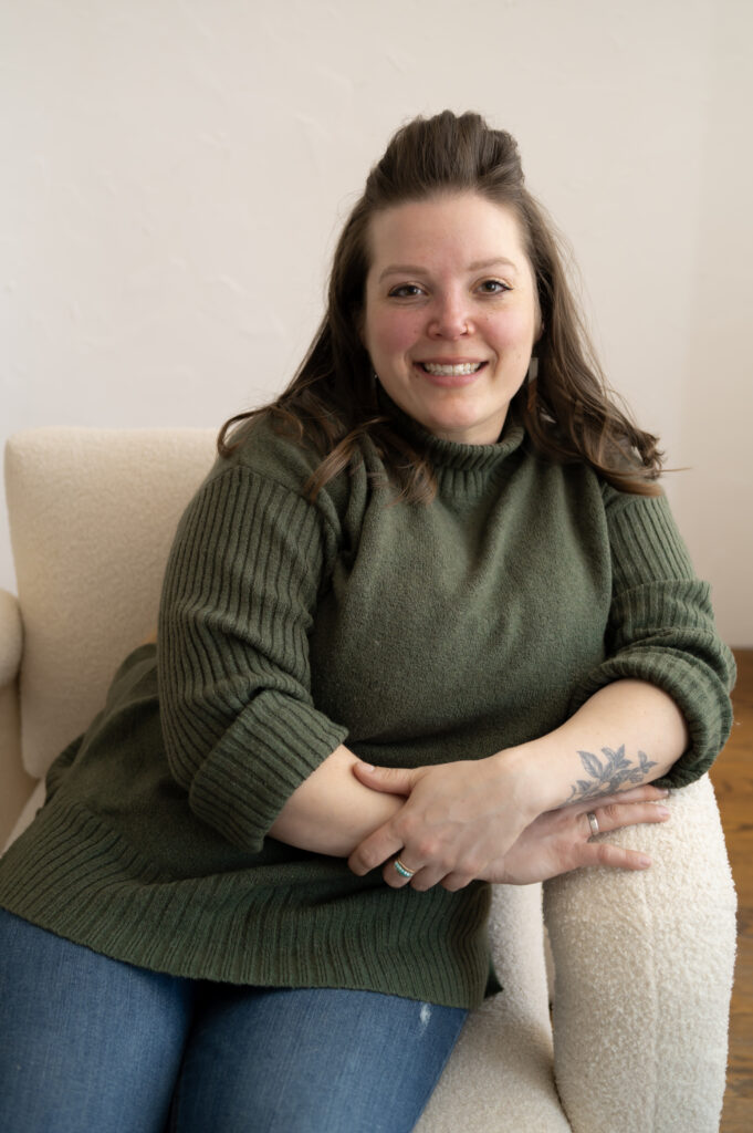 A relaxed, seated portrait of a chef wearing an olive green sweater, dark jeans, and her hair softly resting on her shoulders as she sits in a boucle off-white chair.