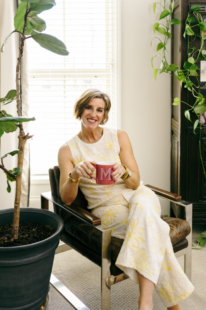 A wardrobe stylist sits in a leather chair next to large plants. She wears a linen set with soft yellow embroidery and holds her favorite red coffee mug.