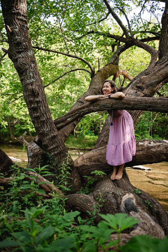 Ana rests her head and arms on a tall horizontal tree branch in front of a creek.