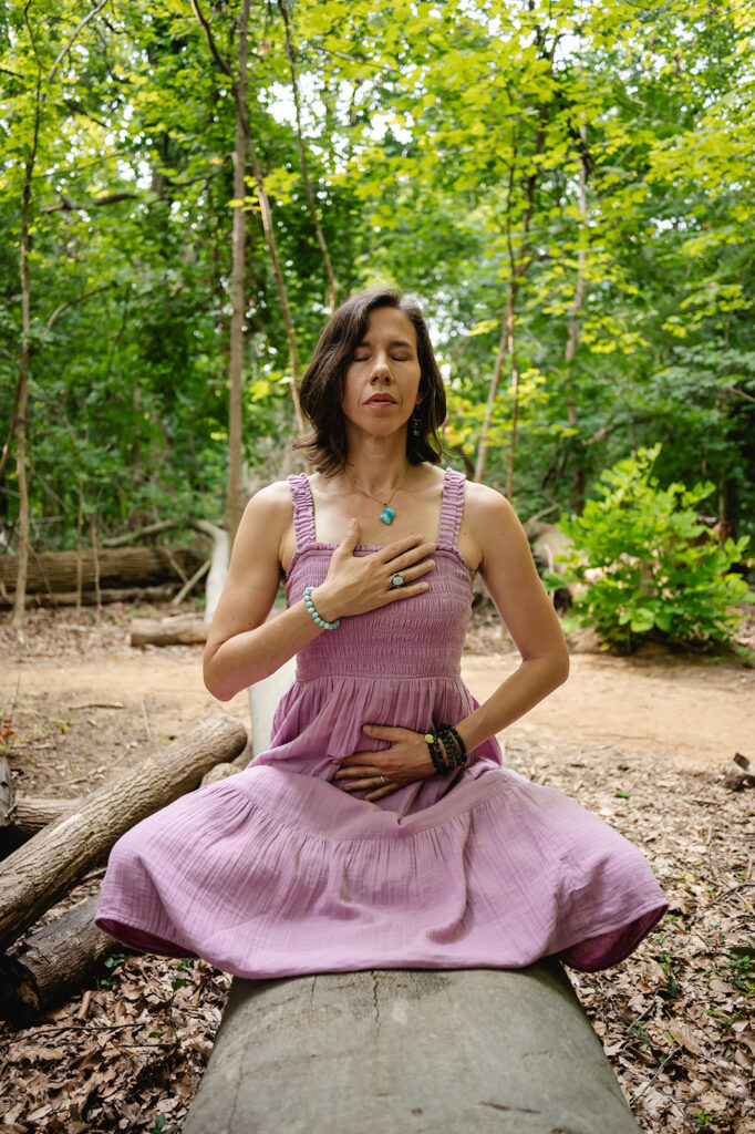 Ana sits atop a fallen log. She has one hand over her heart, the other over her stomach and has both eyes closed in meditation.