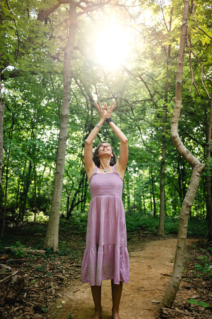 She stands in the middle of the forest path and raises her hands in a crown shape. The sun looks like an orb in the middle of her hands.