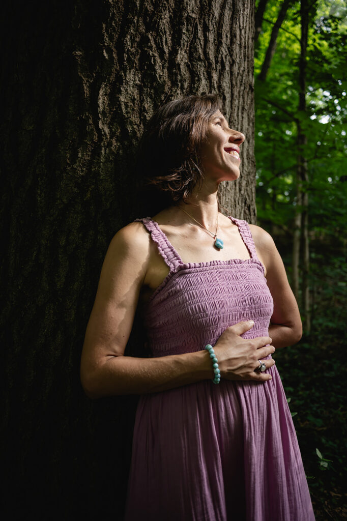 A Dominican woman smiles as she rests against a tree, wearing a purple tank-sleeve dress and a turquoise beaded bracelet.