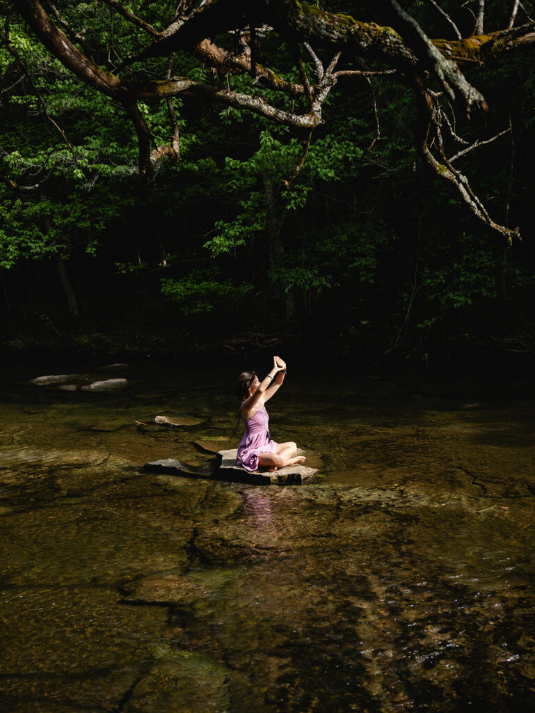 A woman sits on a lone rock in the middle of a forest creek. She holds her hands up in a yoga pose.
