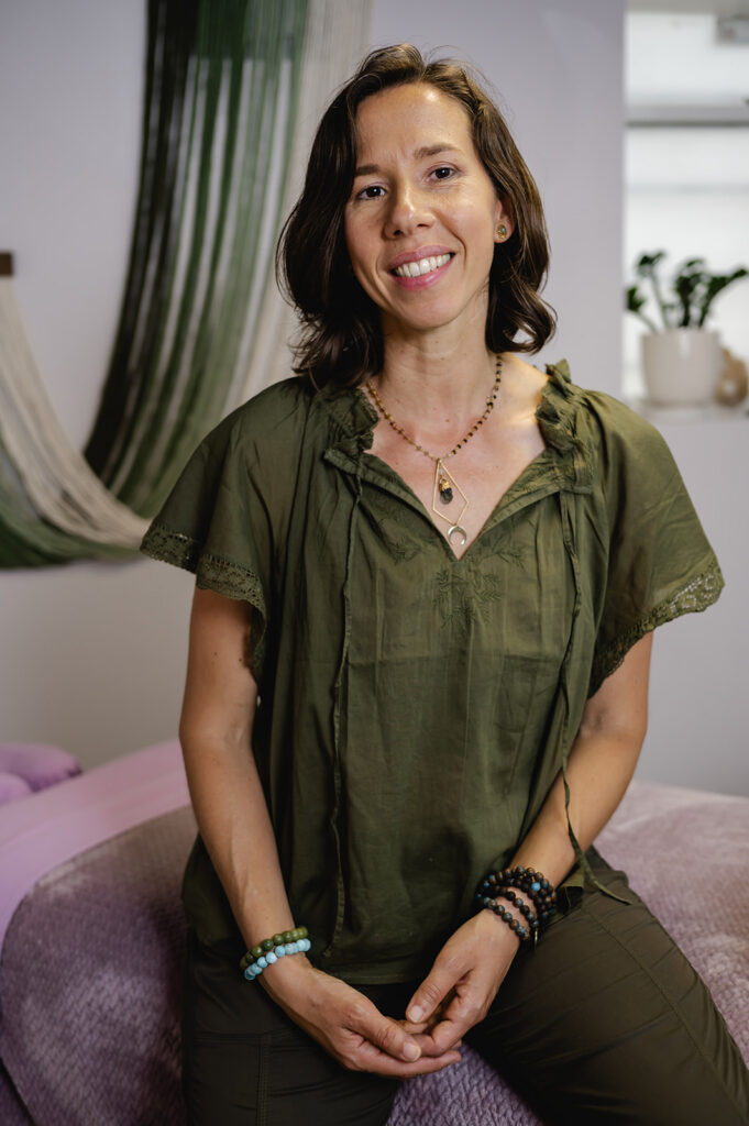 A Dominican woman with shoulder length hair and kind features wears a green cotton top and sits atop a massage bed in her studio.